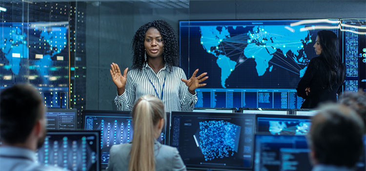 A woman explains information on a screen behind her while those listening look at charts and data on their laptops