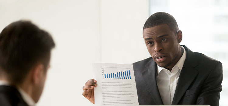 A man at a desk discusses data on paper with another manseks