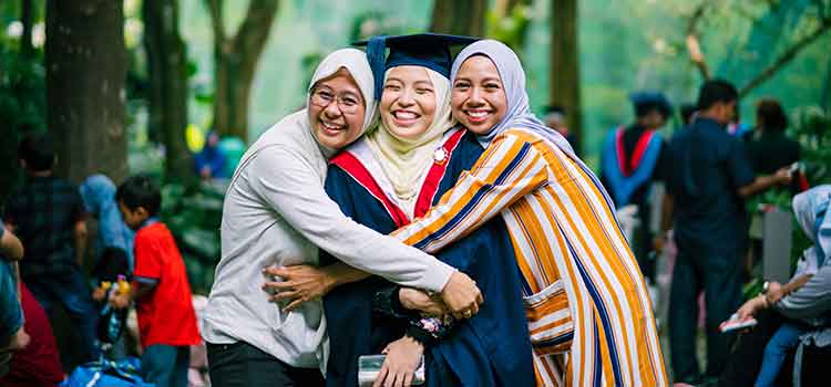 female graduate in cap and gown being hugged by family members