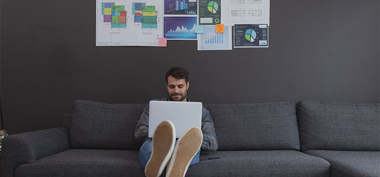 a man sitting on a sofa reads on a laptop with charts on the wall behind him