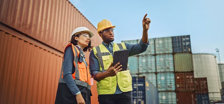 a man in a hardhat standing on a dock among cargo containers gives instructions to a woman in a hardhat